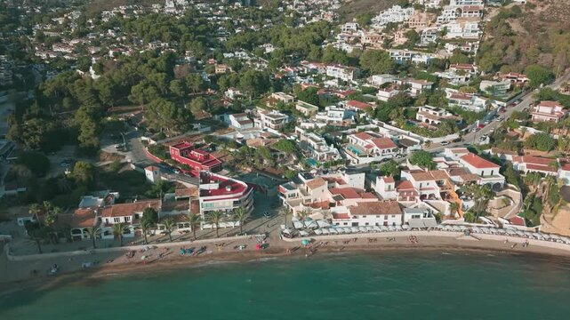 Aerial drone footage of Moraira beach and coastal town in Spain. View of seaside houses, palm trees, and people relaxing on the sandy beach by the Mediterranean Sea.