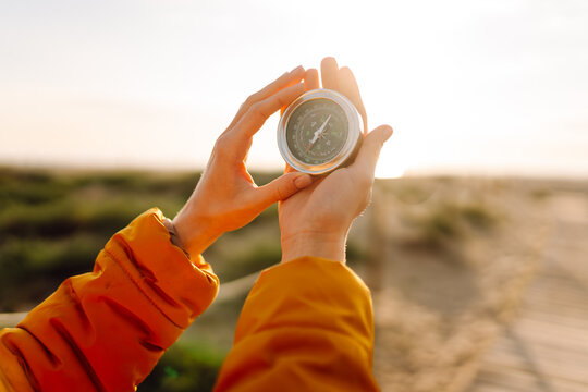A young hiker in a bright jacket and backpack holds a compass and looks out at the seascape. Hiker chooses a direction while standing on a hiking trail at sunset. Concept of adventure, navigation.