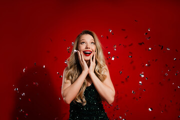 Joyful young woman in green sequin dress celebrates holiday party with confetti against red...