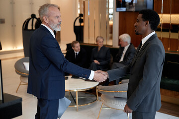 Caucasian middle aged man and Black young adult man shaking hands in modern office lobby, three business professionals sitting in background having discussion, formal business attire