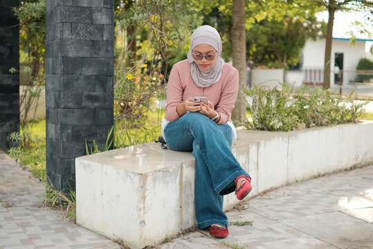 a Muslim woman is sitting in the park while playing with her smartphone - Powered by Adobe