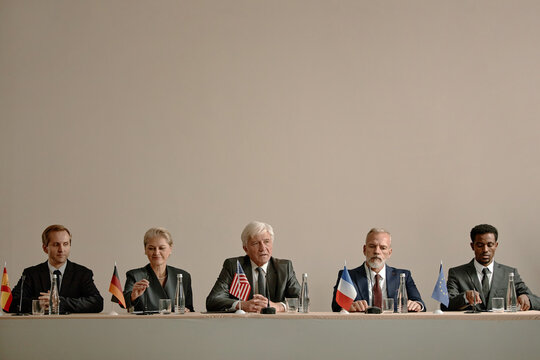Group of middle aged and senior multiethnic men and women sitting at conference table holding microphones, participating in international political meeting with country flags