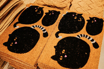 Brown doormats featuring black cats with pink noses and striped tails arranged in a store. The concept conveys playfulness, charm, and cozy domestic warmth