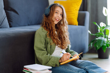 Young woman wearing wireless headphones sits in cozy living room and takes notes in notebook. Beautiful woman enjoys reading book or taking notes. Concept of education, relaxation, and comfort.