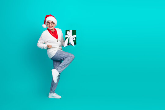 Mature woman in Santa hat holds a green gift against teal background celebrating Christmas and holiday joy