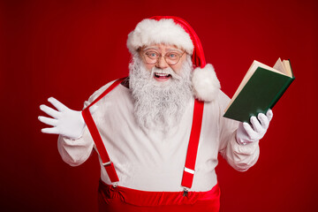 Santa with white beard and red outfit reads a green book while wearing suspenders and glasses in a festive studio set against a bold red background