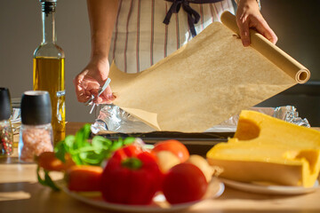 Woman in apron placing baking paper on tray in sunlight kitchen with fresh vegetables, pumpkin, species and olive oil on table. Concept of home cooking, preparation and healthy homemade food