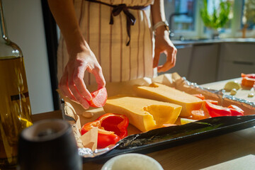 Woman preparing healthy vegetarian food at home, placing tomato on tray with pumpkin and vegetables for roasting. Female chef in apron cooking at kitchen with sunlight