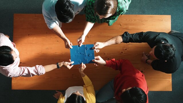 Top down view of skilled business people assemble blue jigsaw puzzle on meeting table. Group of diverse team working together to solve the puzzle. Represented togetherness, cooperative. Convocation.
