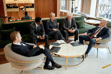 Group of middle aged and senior business professionals including Black man and Caucasian men and woman sitting together in modern office lounge, discussing documents and strategy