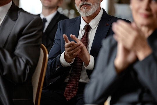 Middle aged Caucasian man in suit sitting among group of professionals clapping hands during formal event, surrounded by other adults in business attire, focus on hands and gesture
