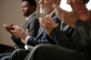Group of middle aged and senior men and women of diverse ethnicities sitting in row clapping hands during event, focus on hands and partial profiles, business attire visible