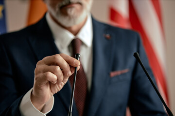 Middle aged Caucasian man adjusting microphone while standing at podium, wearing suit and tie, partial face visible, preparing for public speech with flags in background