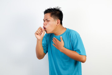 A young Asian man in a blue shirt coughs with his hand in front of his mouth and holds his chest, emphasizing the importance of hygiene and respiratory health.
