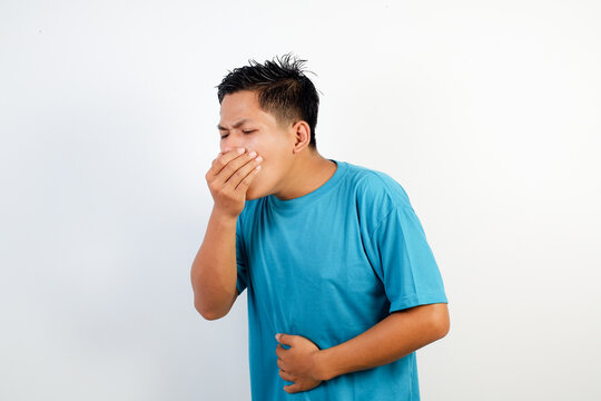 A young Asian man in a blue t-shirt covers his mouth while holding his stomach, showing discomfort from nausea and stomachache. Concept for health and fitness theme.