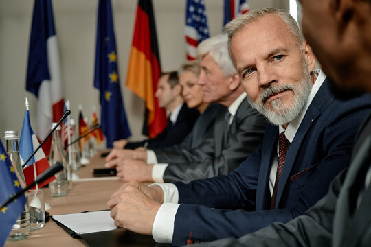 Portrait of middle aged Caucasian man with gray beard sitting at international conference table among diverse group of business professionals discussing global cooperation