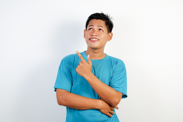 A young Asian man smiling cheerfully, feeling happy and pointing to the side and upward, showing...