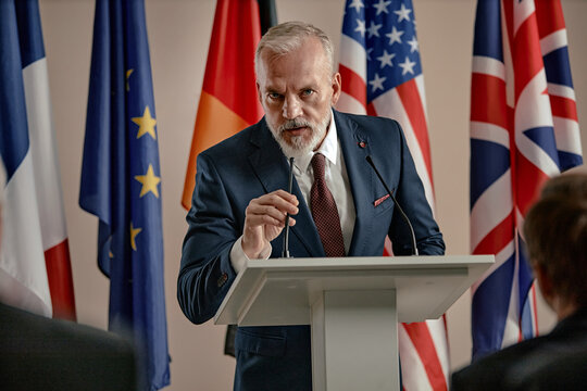Caucasian senior man speaking at podium gesturing with hand during press conference with international flags in background addressing audience in formal setting