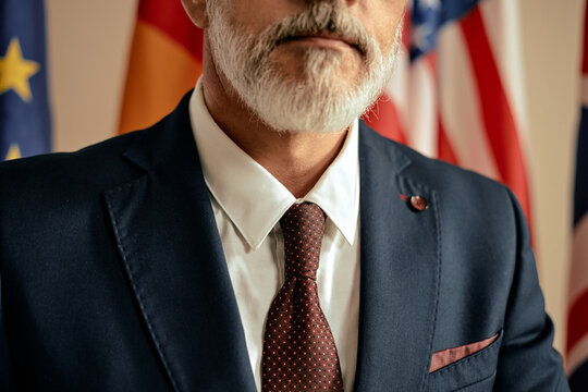 Middle aged man wearing suit and tie standing in front of international flags, showing partial face and upper body, displaying formal appearance and professional demeanor