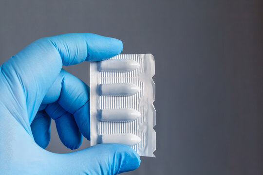 Hand in blue medical glove holding hemorrhoid suppositories in packaging on dark background, close-up.