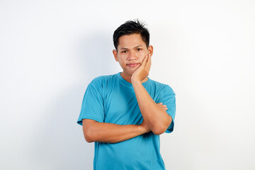 A young Asian man in a blue t-shirt stands against a white background, thoughtfully resting his chin on his hand.