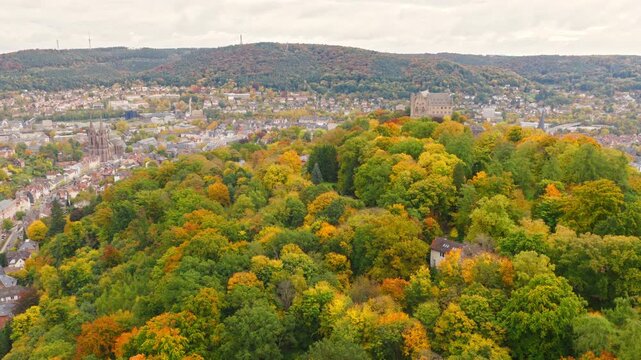 The drone flies over Marburg, showing the old town and the iconic Elisabeth Church, surrounded by trees and the forested hills of the region.