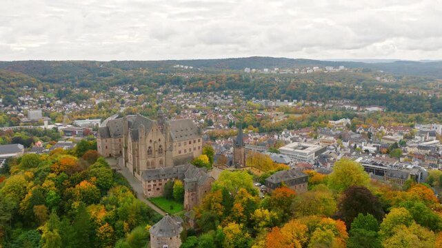 The drone flies over Marburg, showing the old town and the iconic Elisabeth Church, surrounded by trees and the forested hills of the region.
