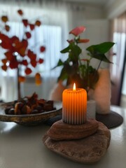 Warm candle flame glowing in a serene autumn still life, surrounded by stones, foliage and soft daylight in a peaceful home setting.
