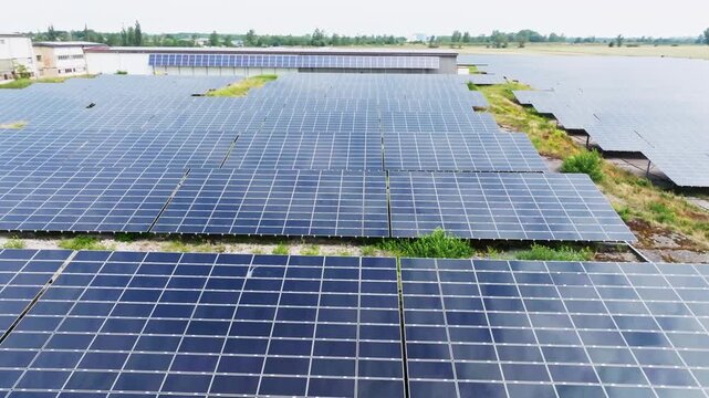 The drone flies over a solar field from a top-down view. Evenly arranged solar panels create a geometric pattern across the green area.