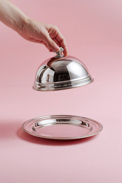 A human hand lifting a shiny silver cloche above an empty metal plate. Minimalist studio setup with a soft pink background. Concept of service, presentation, hospitality, restaurant reveal or surprise