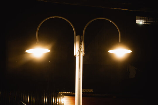 Foto lampu jalan bergaya minimalis dengan dua kap lampu berwarna kuning hangat yang menyala di malam hari, menciptakan suasana tenang dan sunyi. Cahaya lampu menerangi area sekitar dengan nuansa vinta