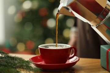 Pouring aromatic coffee from moka pot into cup at wooden table with fir tree branches against...