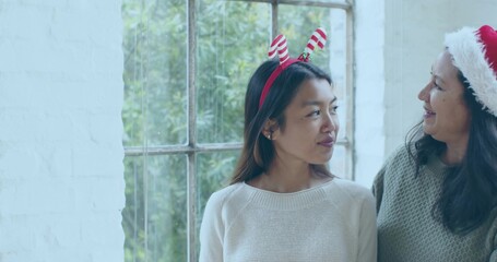 Sharing mother and daughter exchanging warm smiles at window, with knit sweaters and festive hats