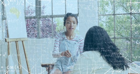 Holding paintbrush, Asian teacher demonstrating painting in studio, wearing overalls, math overlay