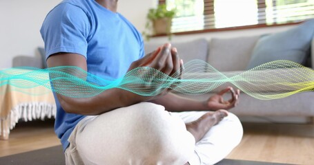 Meditating man wearing blue t-shirt, gray sweats crosslegged on yoga mat at home, with digital wave