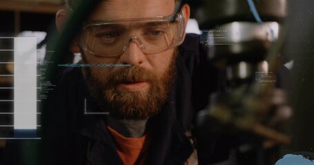 Bearded machinist inspecting spindle and chuck in workshop, wearing goggles, orange shirt with HUD