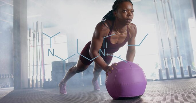 Performing woman wearing maroon tank and shorts holding core while pushing purple med ball at gym - Powered by Adobe