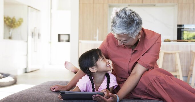 Smiling senior woman in rust dress leaning toward grandchild on ottoman in kitchen holding tablet