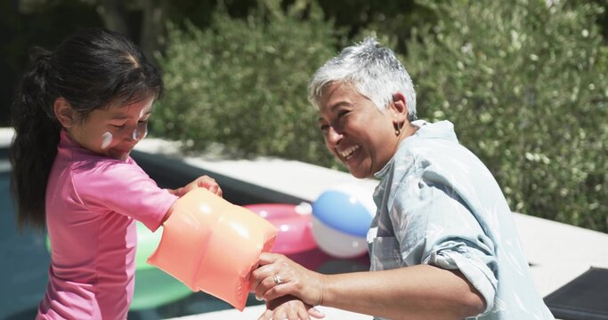 Leaning senior woman in blue shirt reaching child in pink swimsuit at pool holding orange float