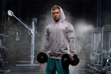 athletic individual in a gray hoodie lifts dumbbells while focusing on bicep curls in a gym setting. The environment features workout equipment and a misty atmosphere.
