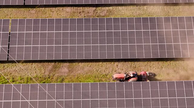 A lawn tractor cuts the grass between solar panels,drone flies over a large solar farm near a forest. Wide landscape
