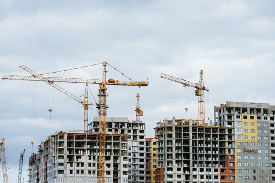 Construction site with several yellow tower cranes actively building multiple unfinished grey residential apartment complexes. Showcasing urban expansion and ongoing development under an overcast sky