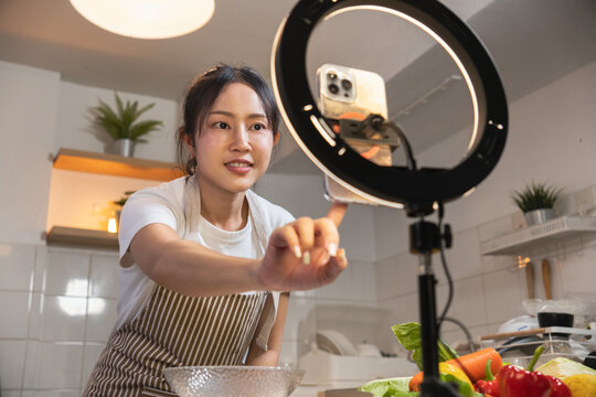Asian woman cooking and recording a video using a smartphone on a ring light for a food vlog or livestream