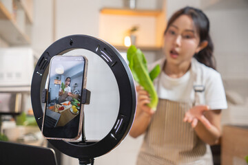 A young woman holding vegetables in the kitchen is talking to a camera phone attached to a ring light. Asian woman live streaming a cooking session