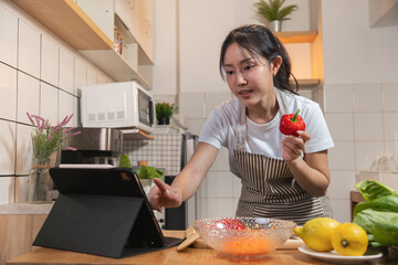 Asian woman teaching an online cooking class, showing a bell pepper while touching a tablet screen