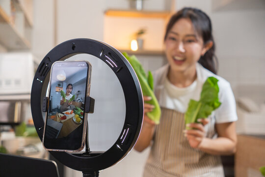 A young woman holding vegetables in the kitchen is talking to a camera phone attached to a ring light. Asian woman live streaming a cooking session
