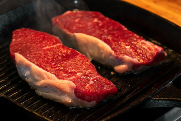 Close-up of Raw Marbled Beef Steaks Searing on Hot Grill Pan. Macro shot highlighting fat rendering, visible steam, and rich red meat color. Ideal for high-protein recipes and culinary content.