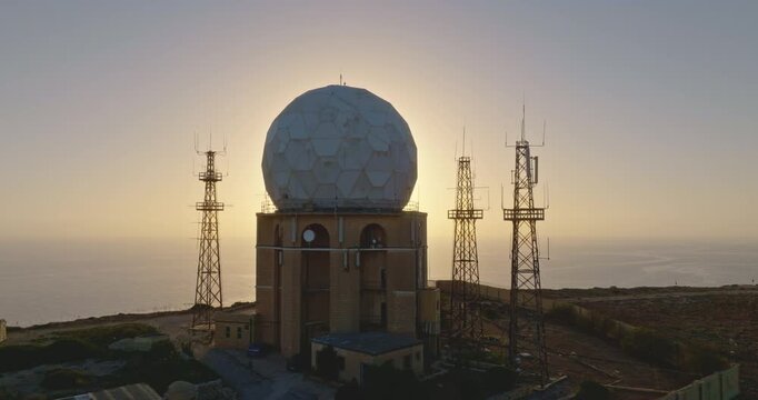 Aerial military ground radio station with radar antenna shelters and radio towers at sunset. The spherical radar dome stands prominently against the serene coastal backdrop. Drone flight footage