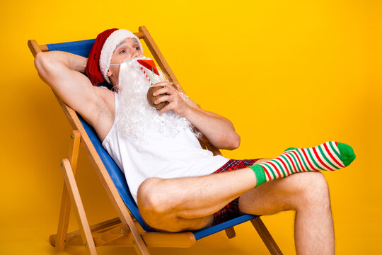 Santa relaxes at home yellow studio wear red hat white beard striped pajama white tank top while sipping mug blue deck chair playful Christmas scene - Powered by Adobe