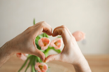 Hands of man making heart over pink tulips at home, close up.